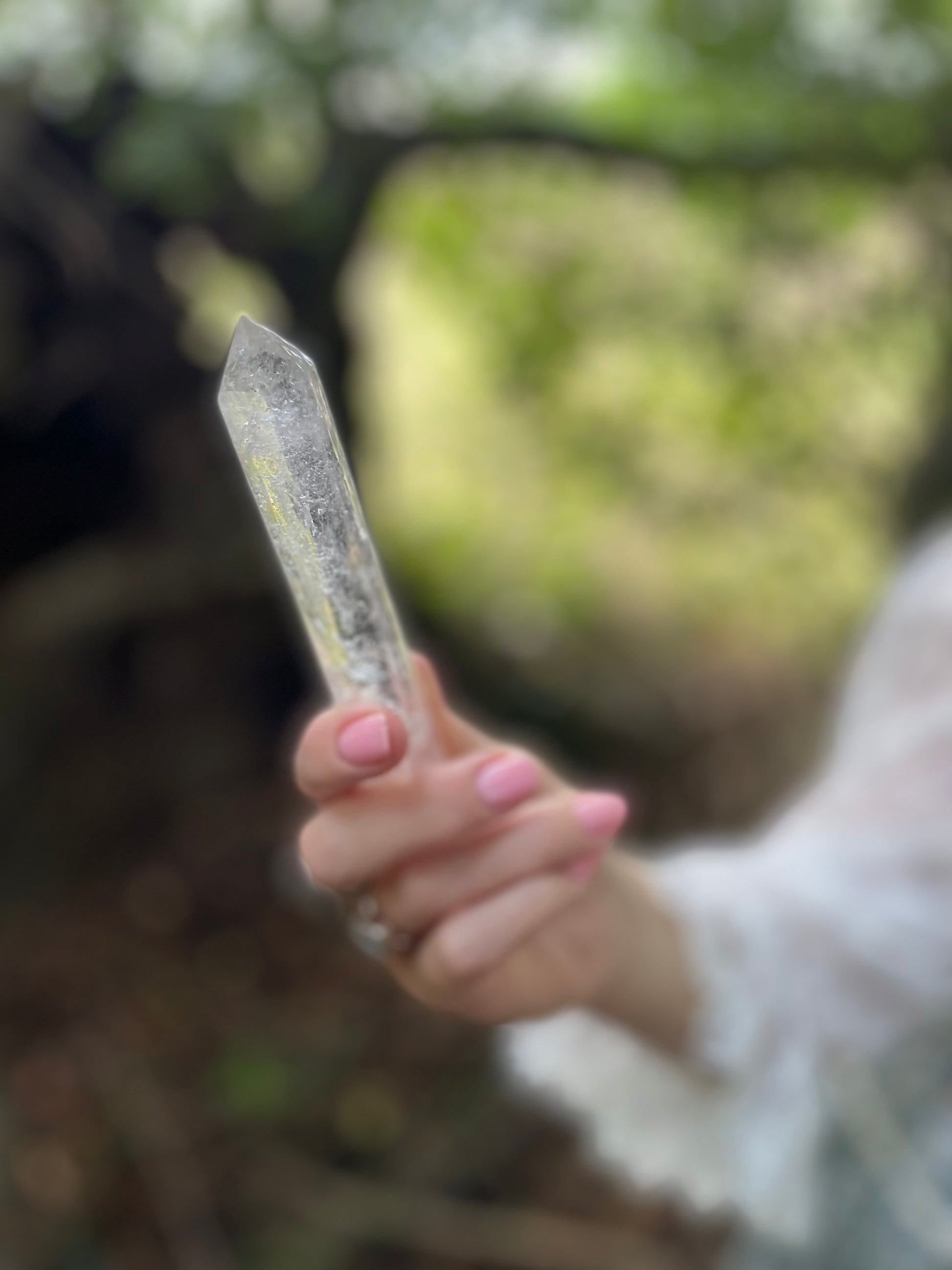 Clear glasslike long crystal wand with inclusions held in models hand in the woods. Model wears a frilly white blouse