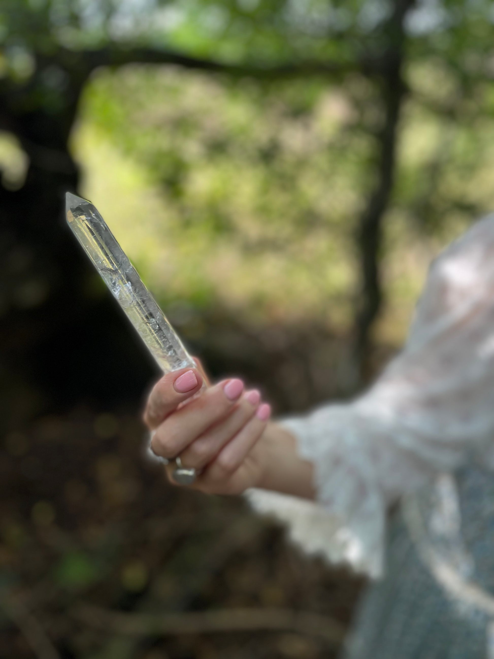 Clear glasslike long crystal wand with inclusions held in models hand in the woods. Model wears a frilly white blouse