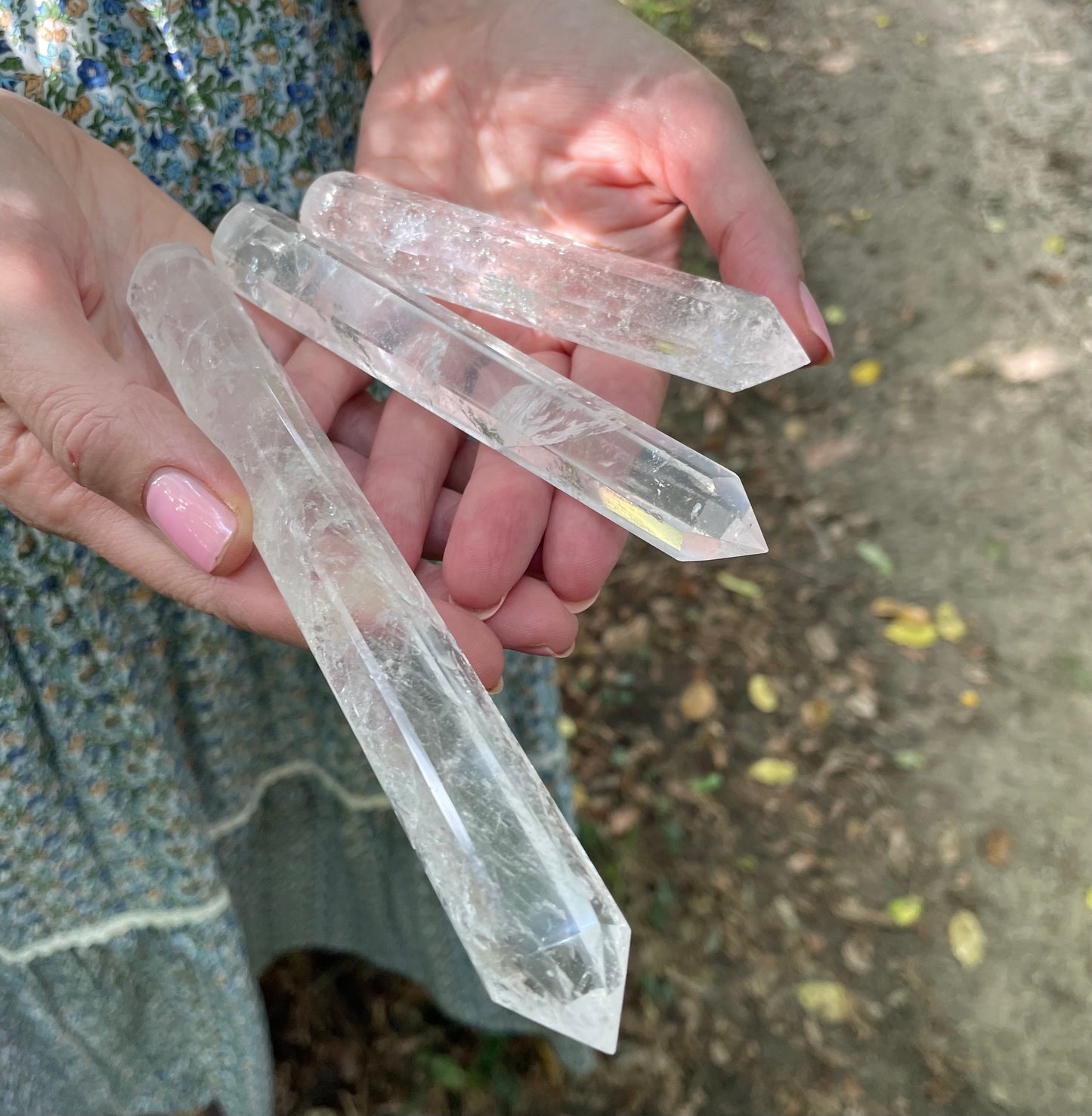 Three clear crystal wands shown in three sizes. Held in models hand showing her blue floral skirt. The background is the forest.