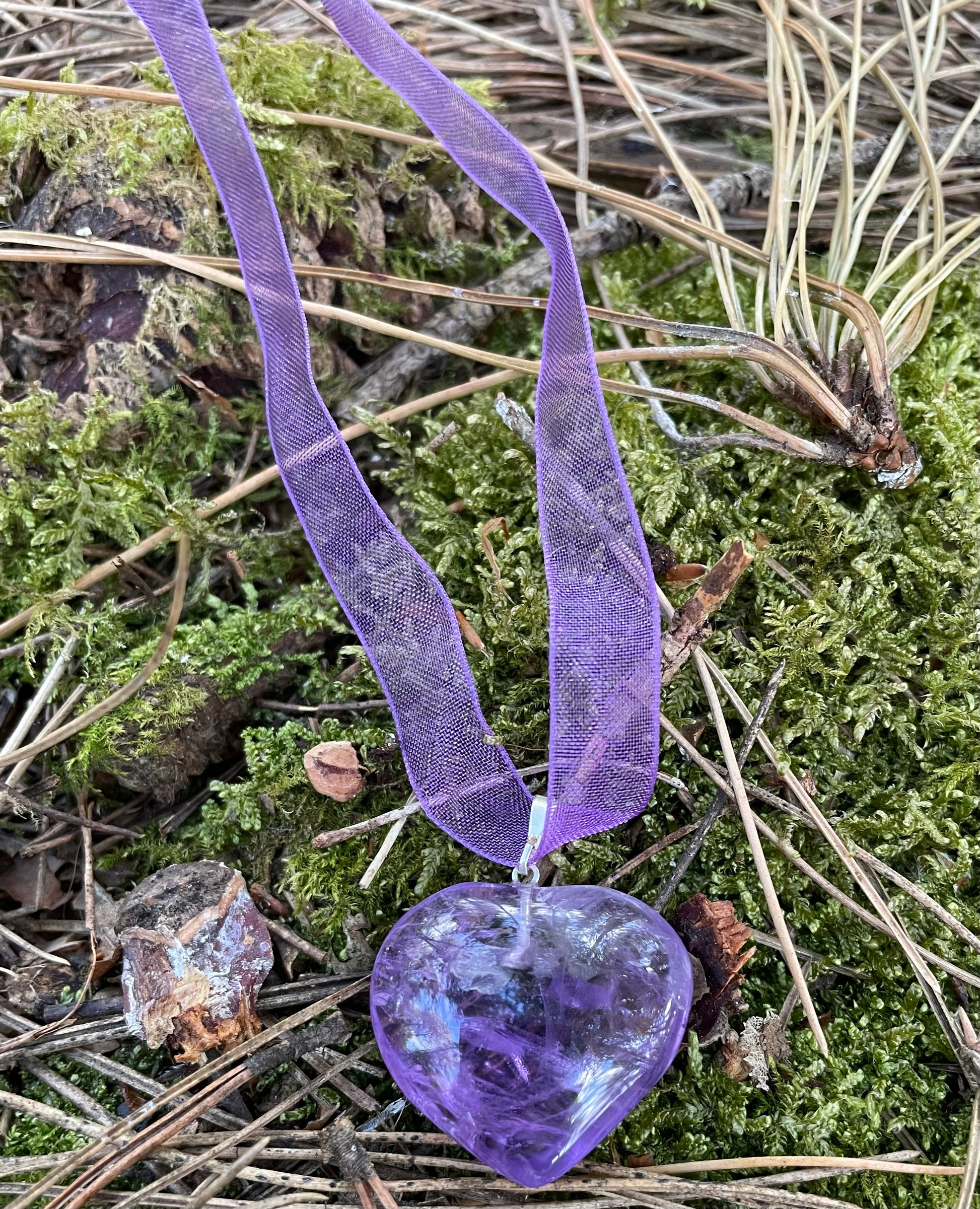 Purple heart shaped Amethyst pendant threaded on purple ribbon. It is photographed in the woods on foliage.