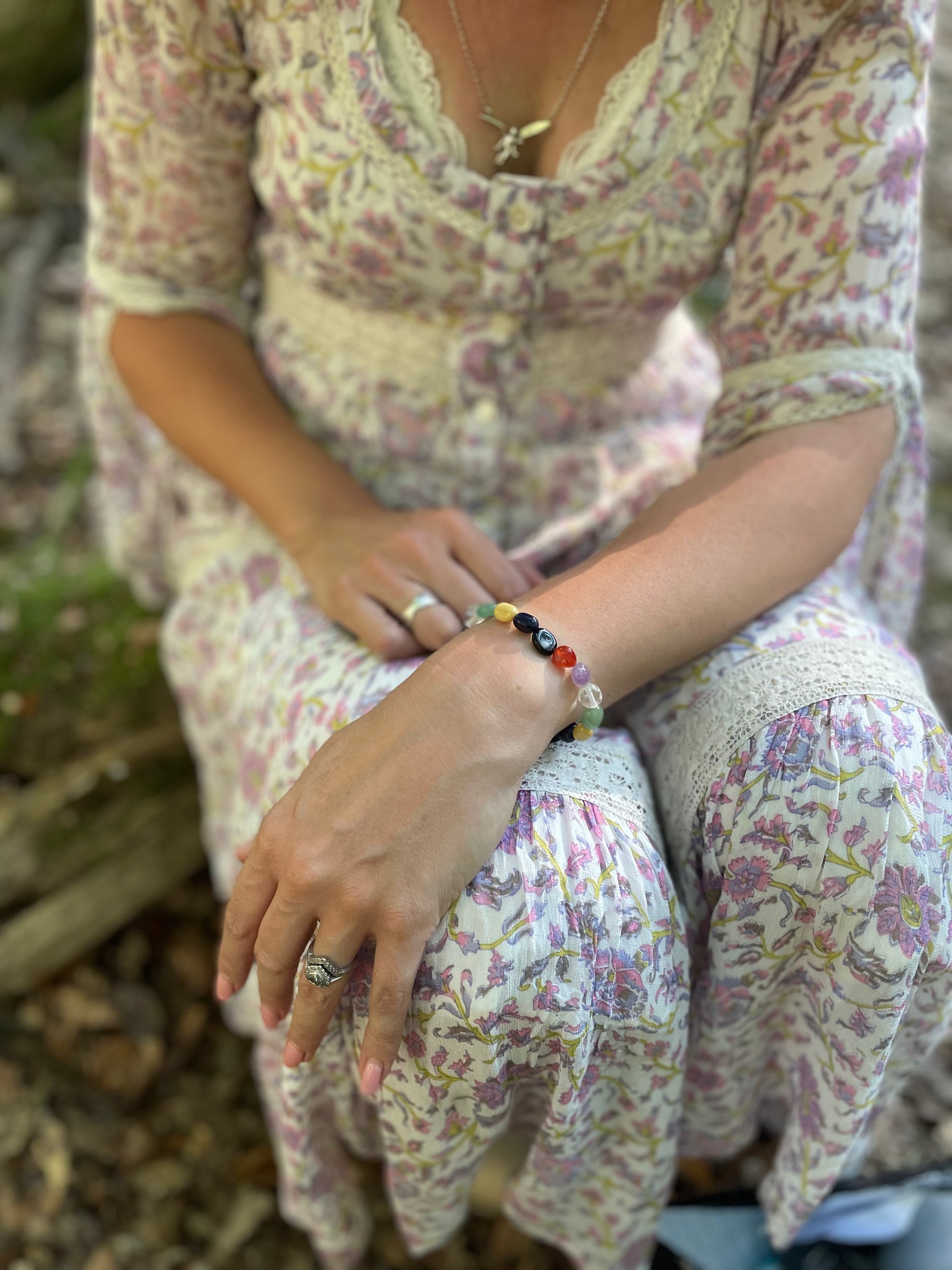 Model wearing multi -coloured tumble stone crystal bracelet. She is sitting in the woods wearing a lilac floral dress.